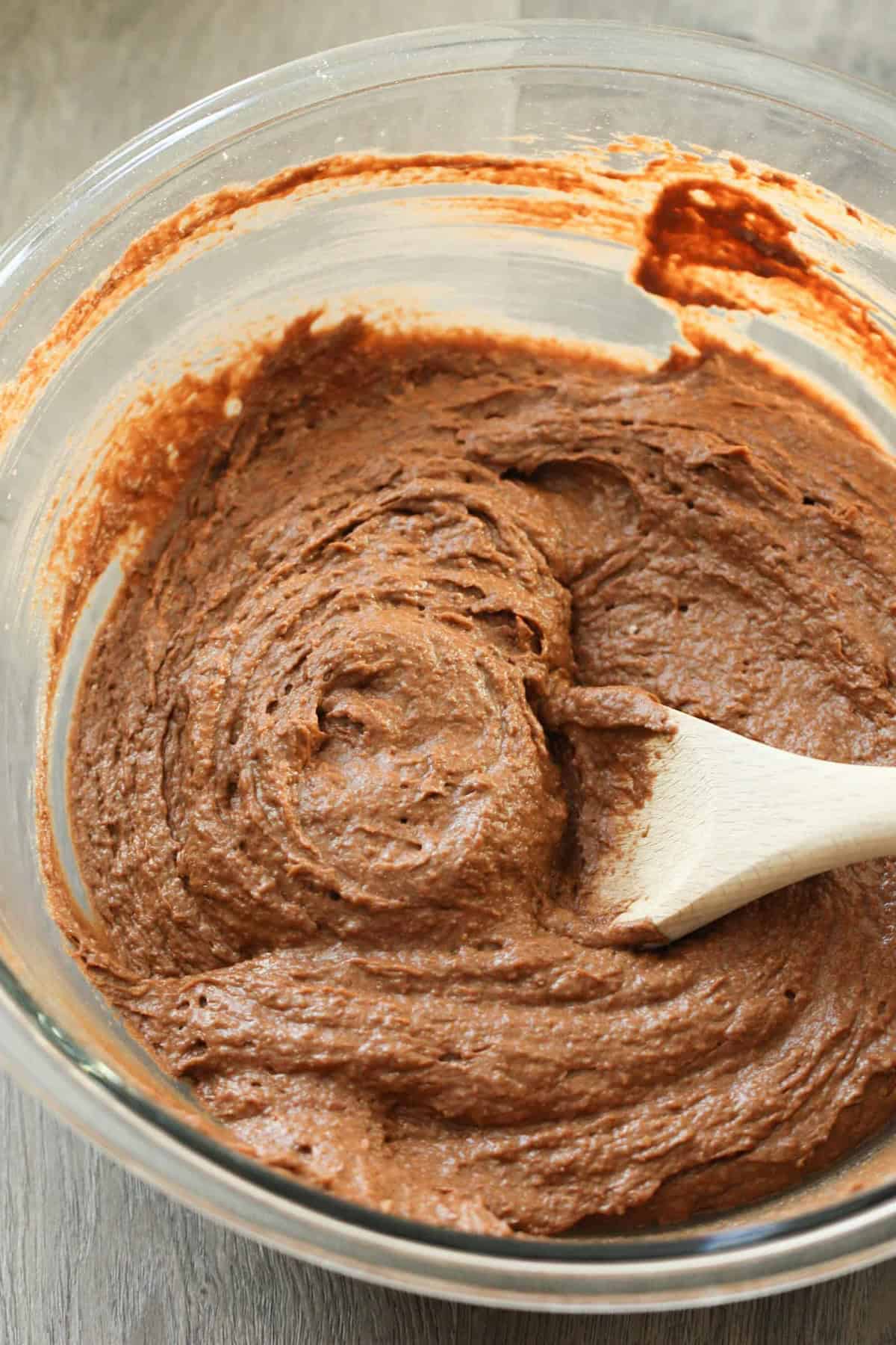 A clear glass bowl filled with chocolate brownie batter is mixed with a wooden spoon.