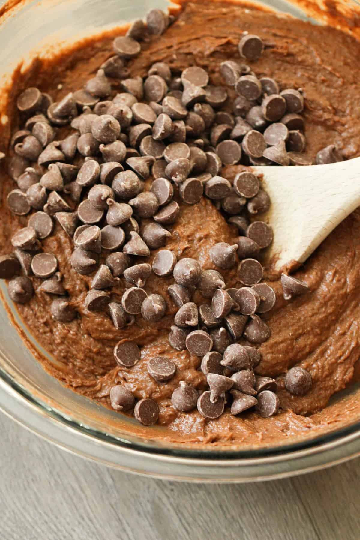 A glass bowl filled with chocolate batter, topped with chocolate chips, being mixed with a wooden spoon.