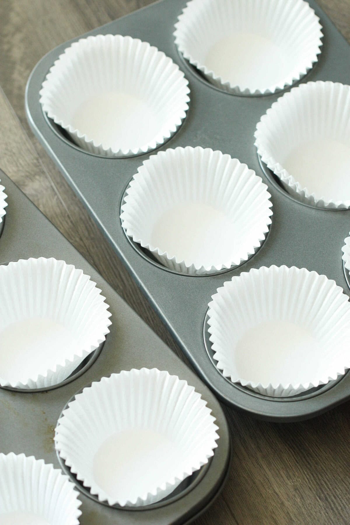 Two muffin trays containing white paper liners are placed side by side on a wooden surface.