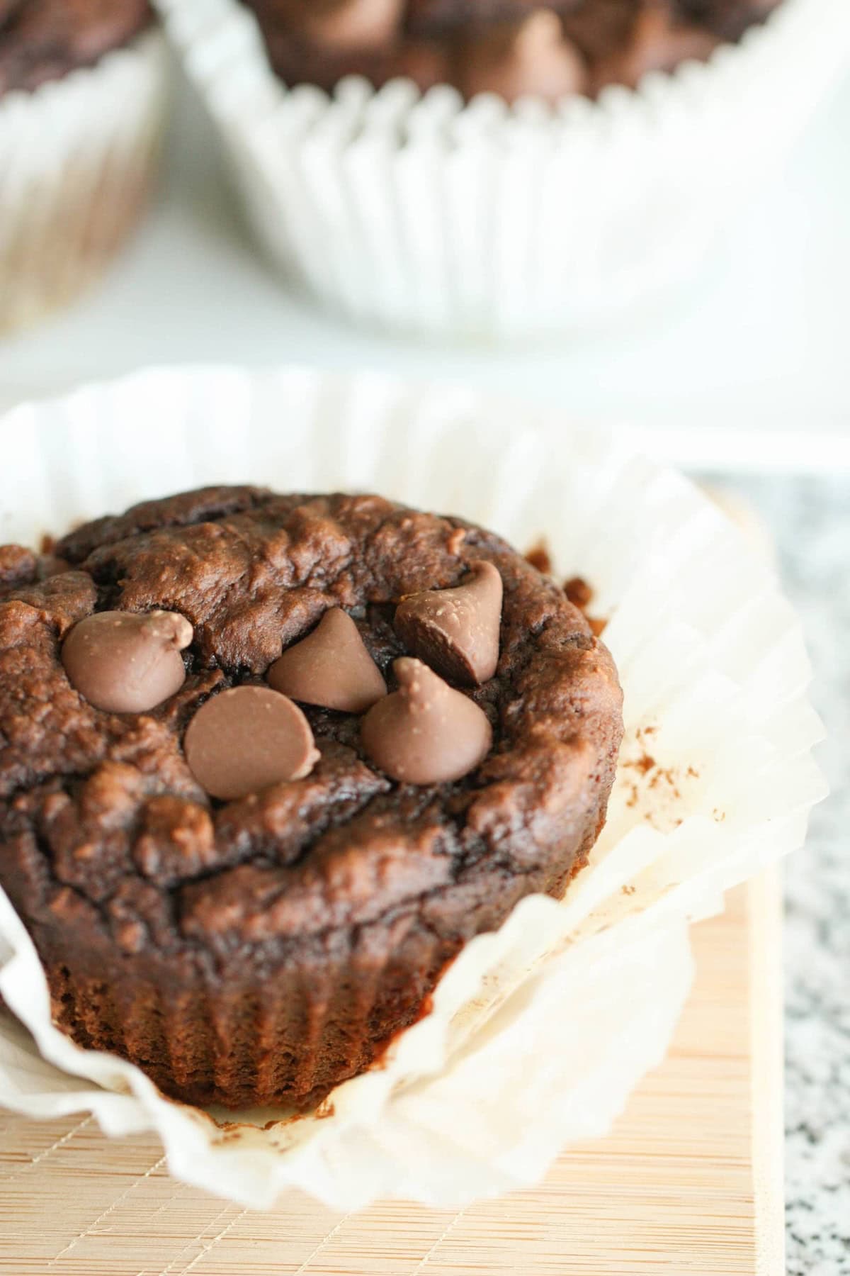 A close-up of a pumpkin chocolate muffin with chocolate chips on top, partially unwrapped from a white paper liner, placed on a light-colored wooden surface.