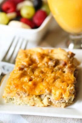 A slice of cheesy casserole on a plate with a fork beside it. A bowl of mixed fruit and a glass of orange juice are in the background.