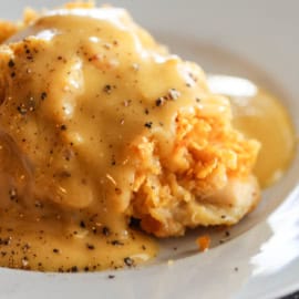 A piece of baked chicken topped with a thick layer of cheese and creamy gravy, garnished with black pepper, served on a white plate.