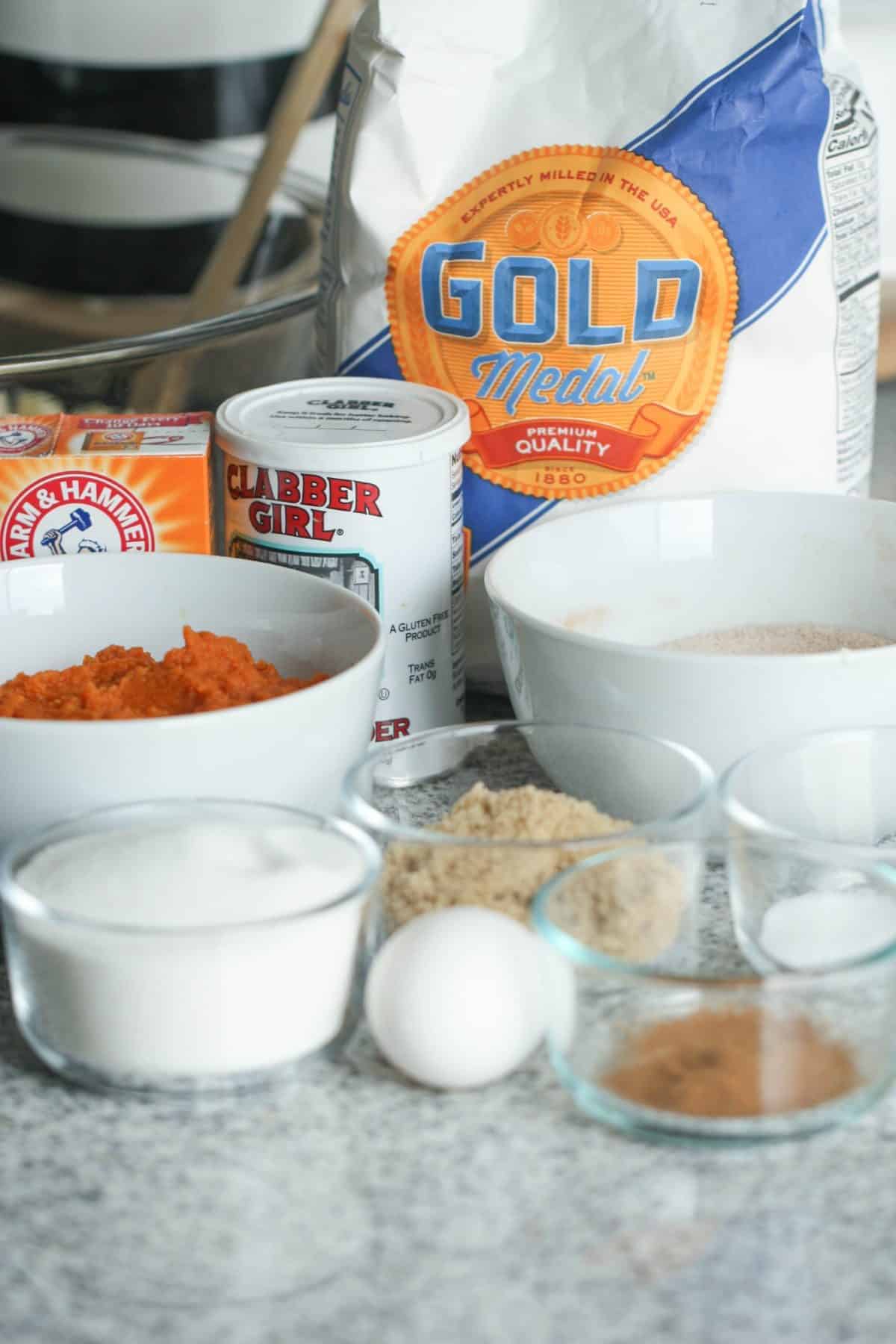 Ingredients for baking, including flour, brown sugar, white sugar, egg, spices, baking powder, baking soda, and pumpkin puree, are arranged on a kitchen counter.