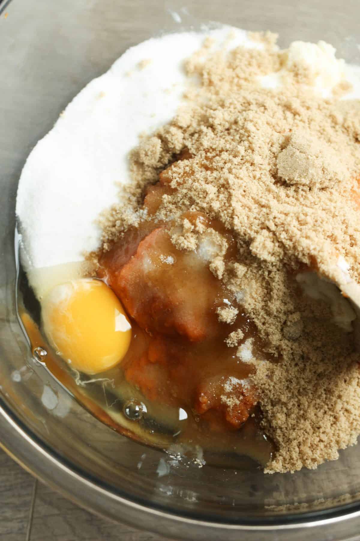 A mixing bowl containing an egg, granulated sugar, brown sugar, and wet ingredients before mixing.