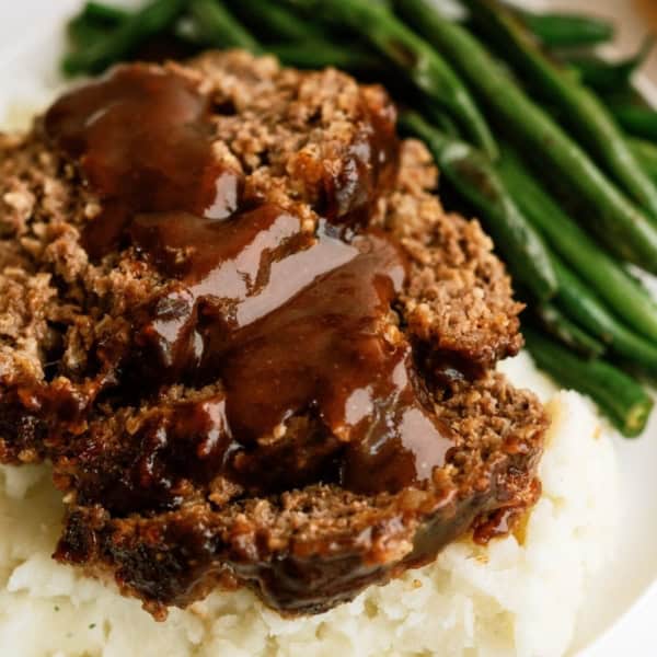 A plate of meatloaf slices topped with gravy, accompanied by mashed potatoes and green beans.