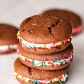 A stack of three chocolate whoopie pies with colorful sprinkles and a cream filling are arranged on a white surface.