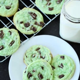 Three green cookies with chocolate chunks on a white plate, with one more green cookie on a cooling rack in the background.