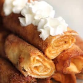 Close-up of a stack of rolled crepes topped with dollops of whipped cream. The crepe filling appears to be a light, spreadable substance. The background is blurred.