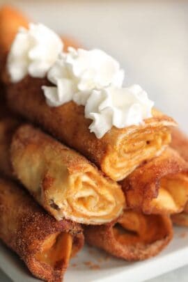 Close-up of a stack of rolled crepes topped with dollops of whipped cream. The crepe filling appears to be a light, spreadable substance. The background is blurred.