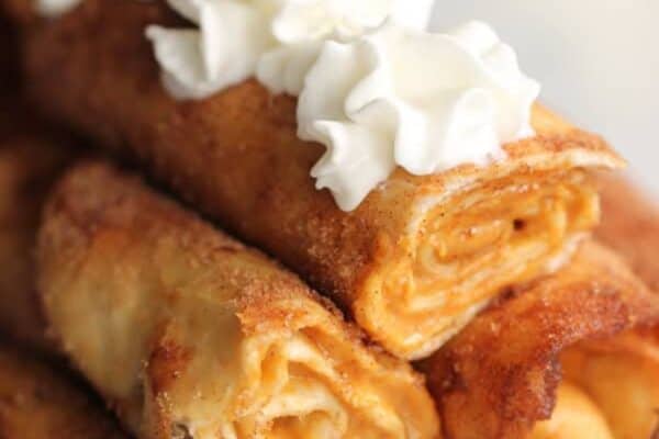 Close-up of a stack of rolled crepes topped with dollops of whipped cream. The crepe filling appears to be a light, spreadable substance. The background is blurred.