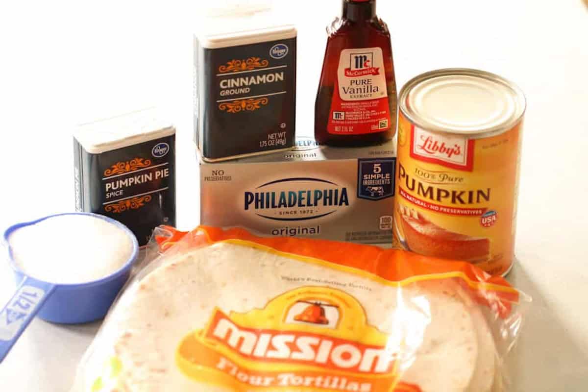 Assorted baking ingredients including canned pumpkin, ground cinnamon, pumpkin pie spice, pure vanilla extract, cream cheese, sugar, and flour tortillas.