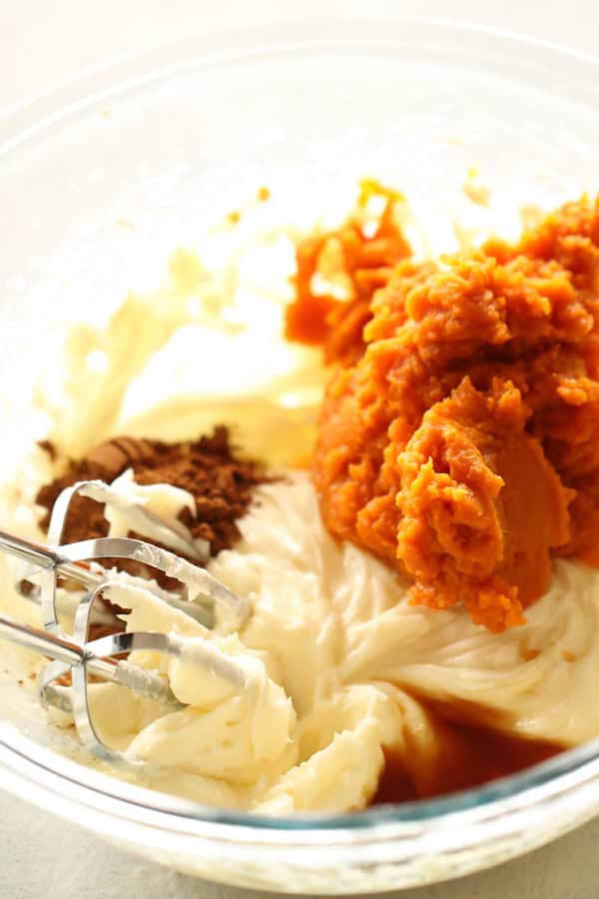 A glass bowl contains a mixture of whipped ingredients including cream cheese, pumpkin puree, and various spices, being mixed with a hand mixer.