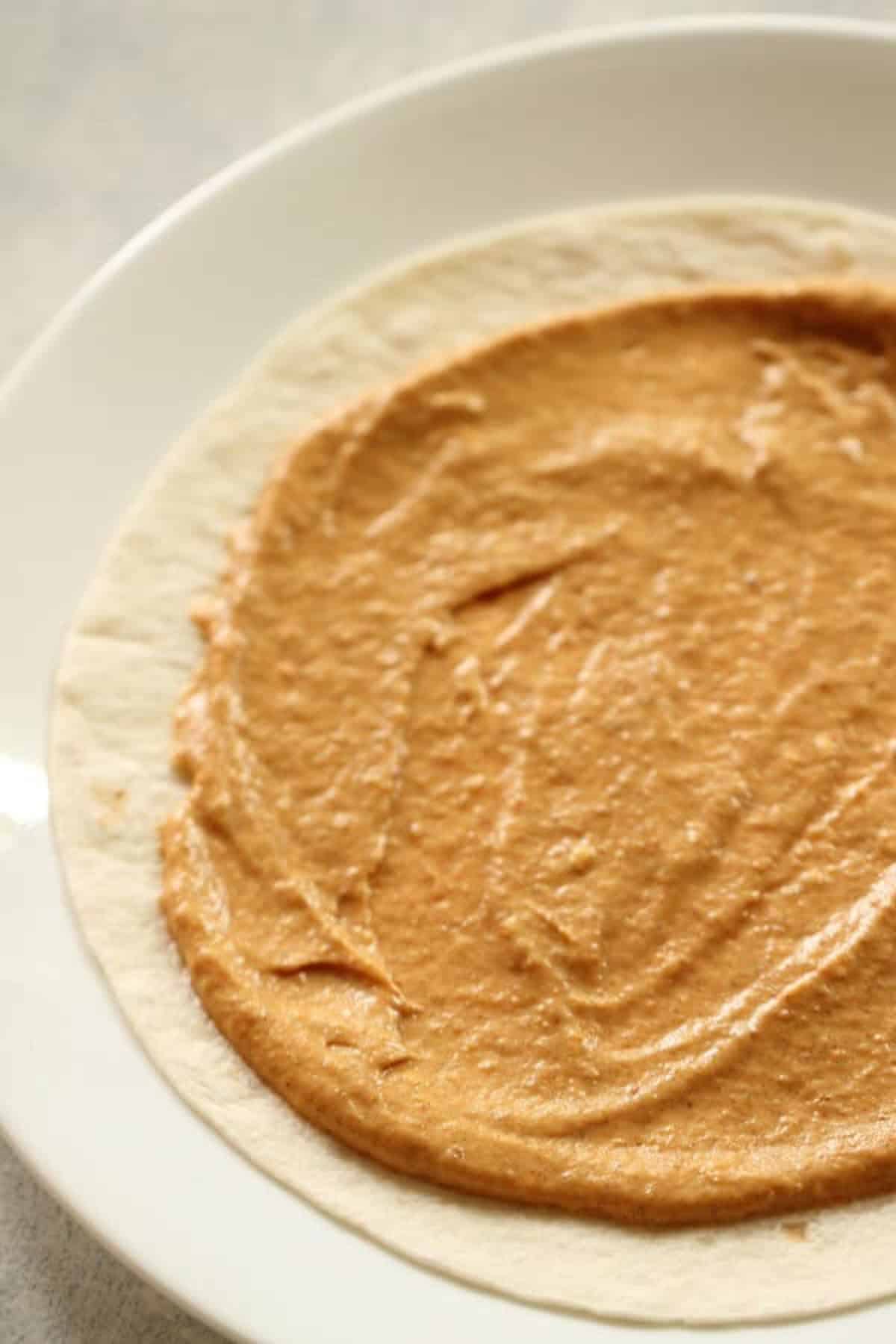 A tortilla spread with a layer of peanut butter is placed on a white plate.