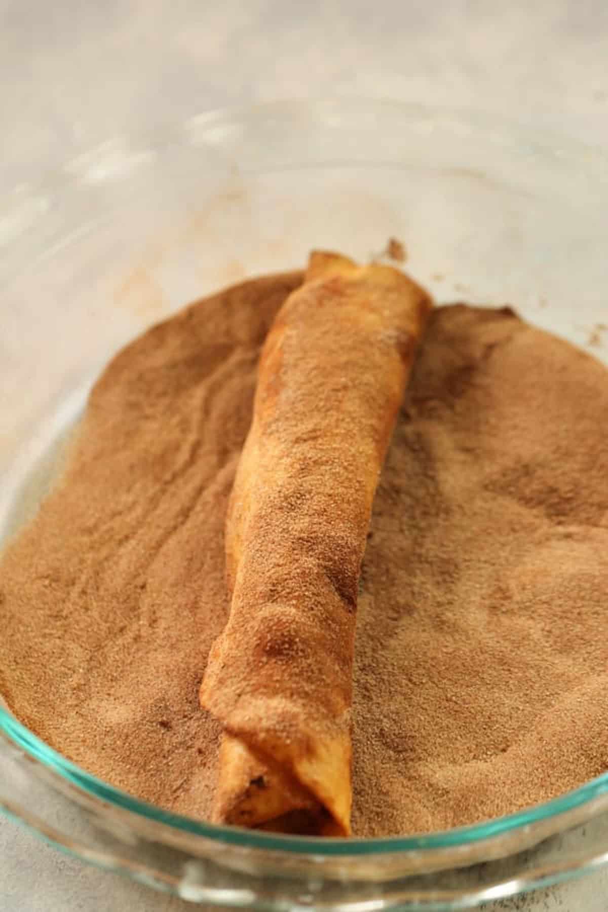 A rolled pastry is coated in cinnamon sugar in a glass dish.