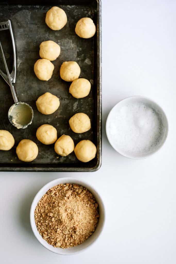 Cookie dough balls on a baking sheet with a small bowl of graham cracker crumbs and a small bowl of foamy egg white.