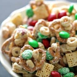 A bowl of snack mix containing pretzels, cereal squares, round cereals, peanuts, and red and green candies.