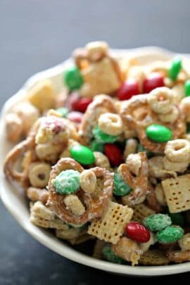 A bowl of snack mix containing pretzels, cereal squares, round cereals, peanuts, and red and green candies.