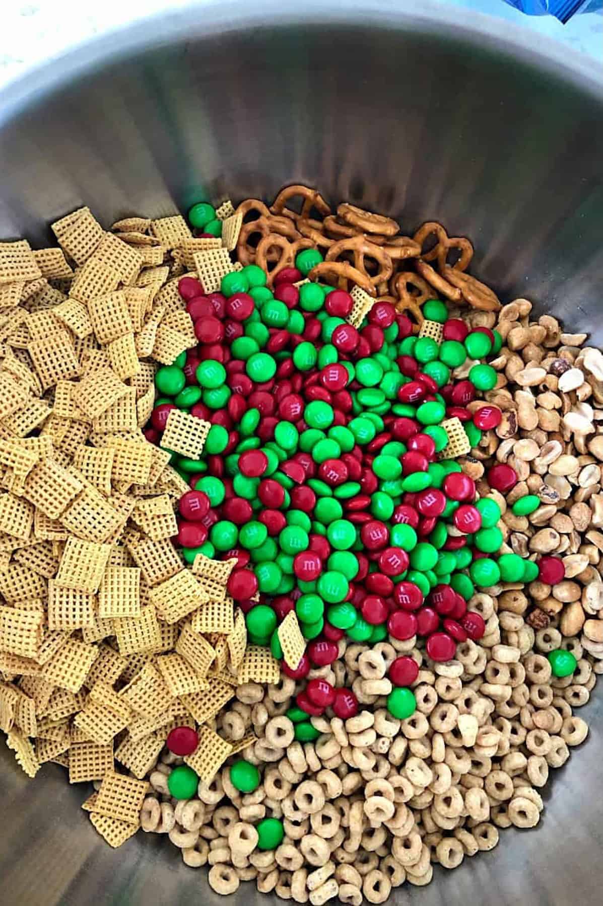 A large stainless steel bowl with a mix of cereal squares, red and green chocolate candies, mini pretzels, and cereal loops.