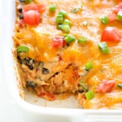 A close-up of a baked casserole with melted cheese, diced tomatoes, and chopped green onions. A portion has been scooped out, revealing a creamy mixture with black beans and other ingredients.