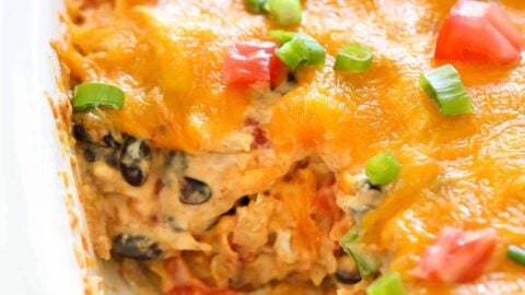 A close-up of a baked casserole with melted cheese, diced tomatoes, and chopped green onions. A portion has been scooped out, revealing a creamy mixture with black beans and other ingredients.