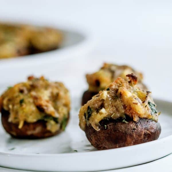 Three stuffed mushrooms filled with a savory mixture are placed on a white plate, with more stuffed mushrooms visible in the background.