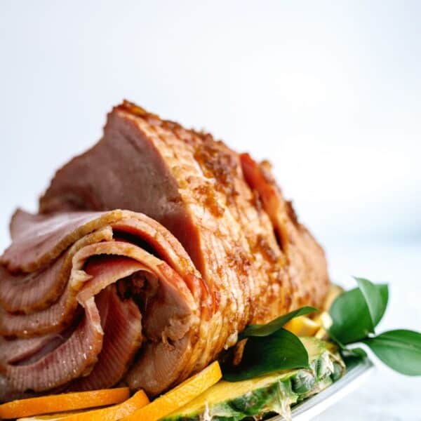 A cooked ham with crispy edges is displayed on a white plate, garnished with green leaves and slices of yellow fruit.