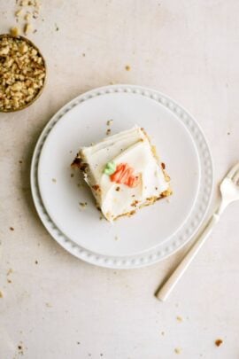 A slice of frosted cake with a small flower decoration on a white plate, accompanied by a fork and a small bowl of crushed nuts.