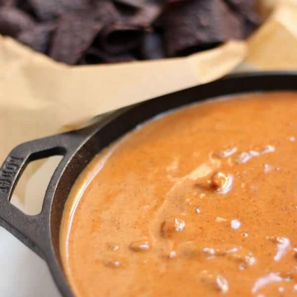 A close-up of a cast iron skillet filled with melted cheese dip. In the background, there is a paper-lined basket containing dark-colored tortilla chips.