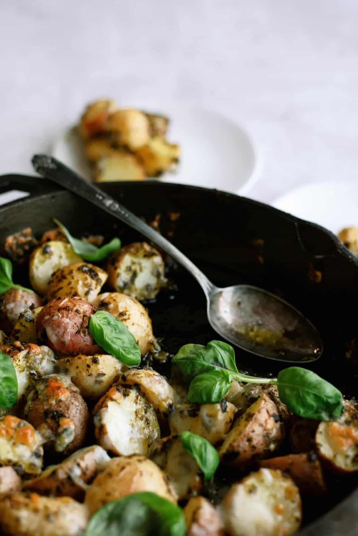 A skillet filled with roasted potatoes topped with melted cheese and garnished with basil leaves. A spoon rests in the skillet. In the background, a plate holds more potatoes.