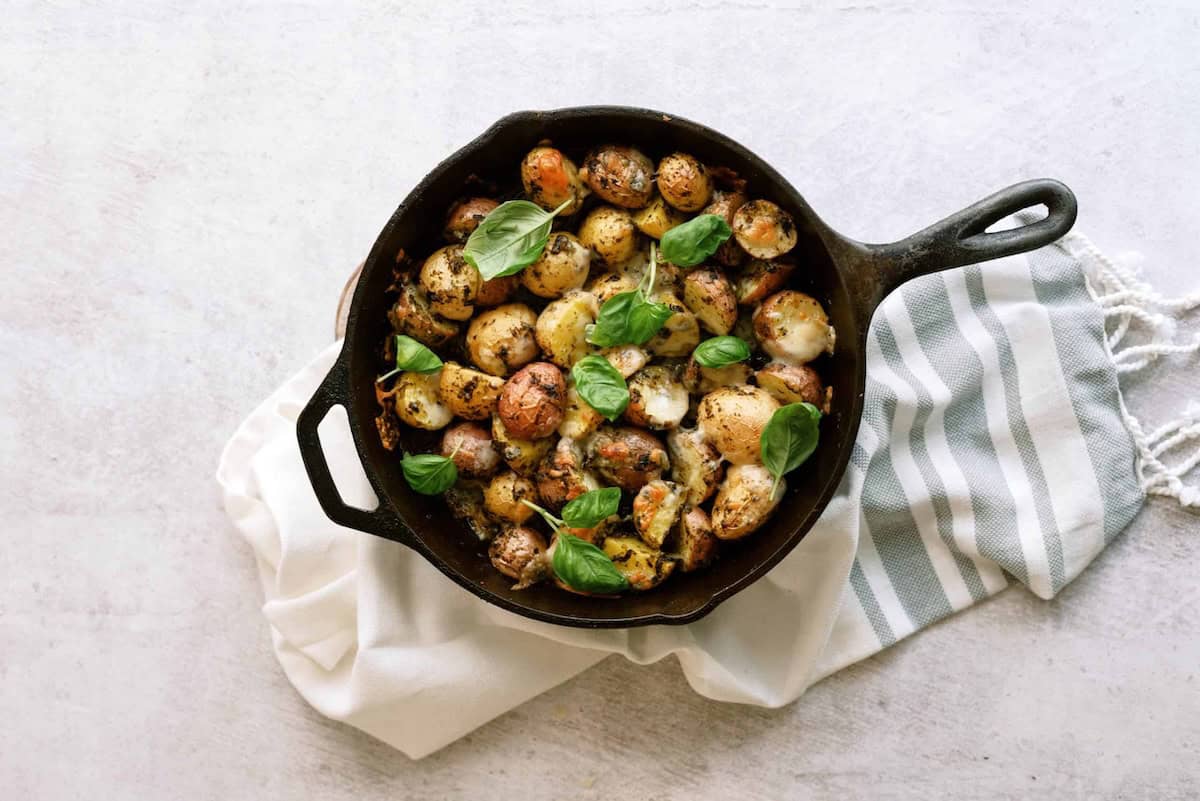 A cast iron skillet with roasted potatoes and fresh basil leaves on a white cloth with gray stripes.