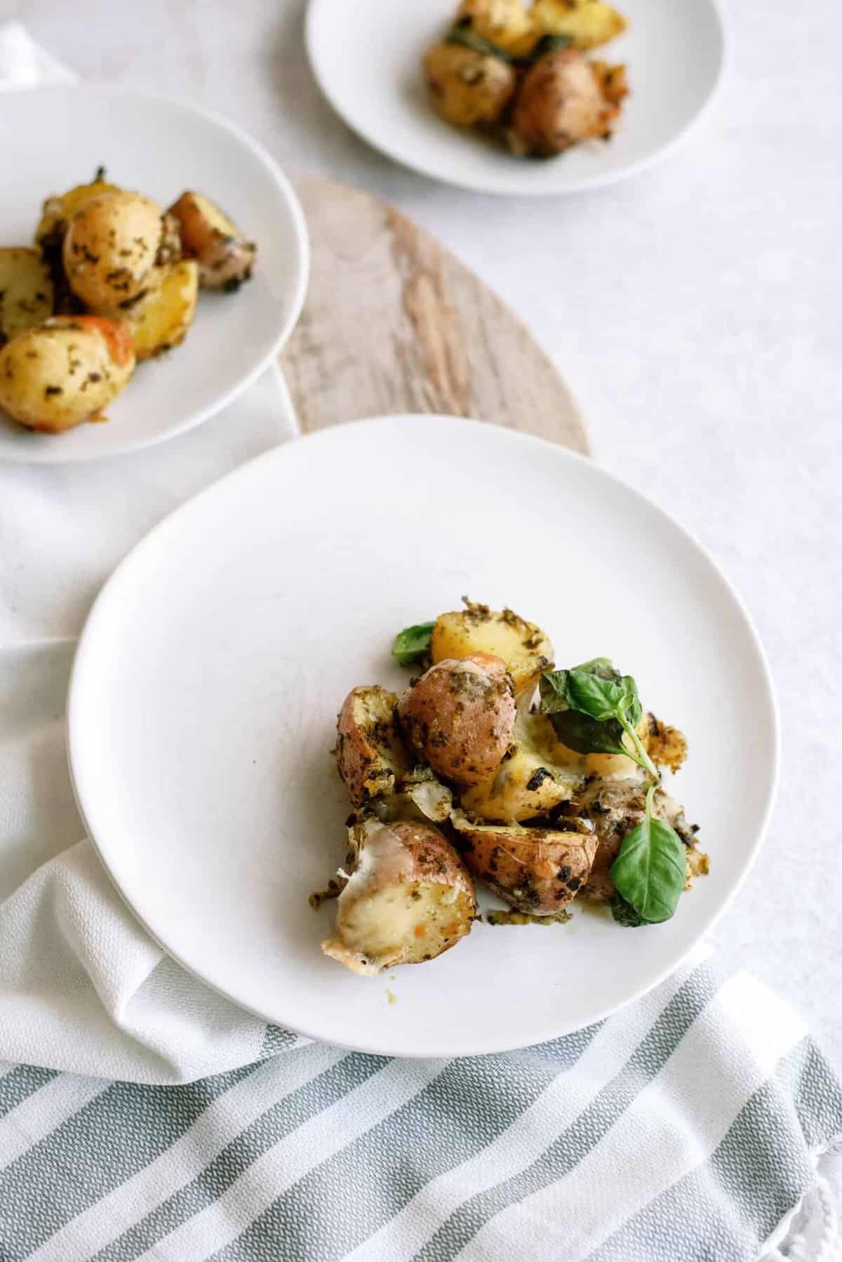 Plates of roasted new potatoes garnished with herbs and a basil leaf on a white table with a striped cloth.