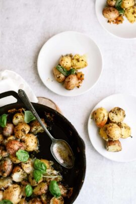 A skillet with roasted potatoes garnished with herbs. Three white plates, each with a serving of the potatoes, are arranged nearby. A spoon rests in the skillet.