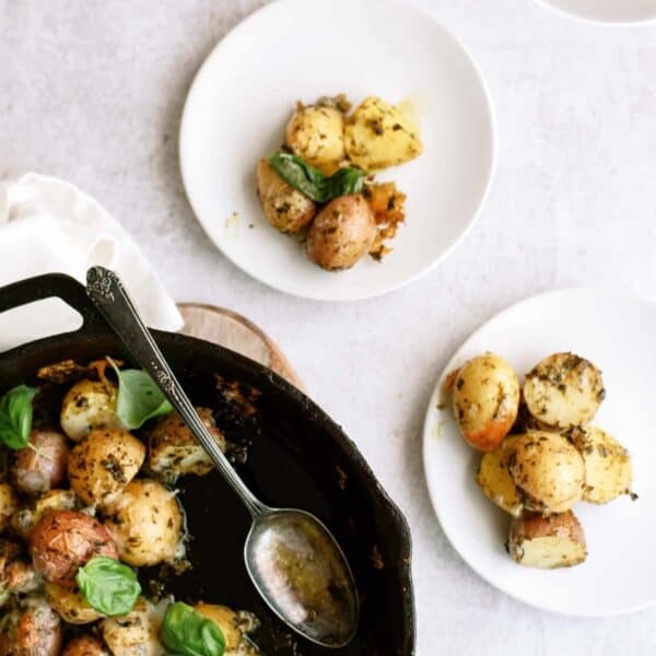 A skillet with roasted potatoes garnished with herbs. Three white plates, each with a serving of the potatoes, are arranged nearby. A spoon rests in the skillet.