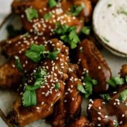 A plate of glazed chicken wings garnished with sesame seeds and cilantro, next to a small dish of dipping sauce.