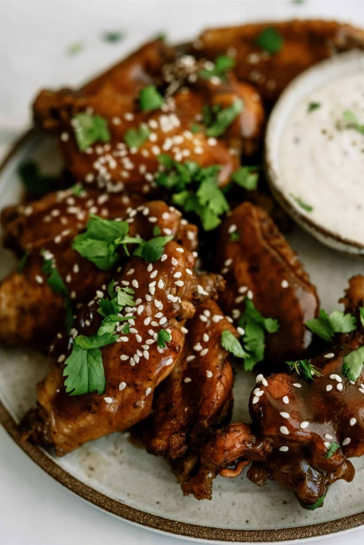 A plate of glazed chicken wings garnished with sesame seeds and cilantro, next to a small dish of dipping sauce.