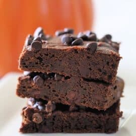 A stack of three pumpkin brownies on a white plate, with an orange pumpkin blurred in the background.