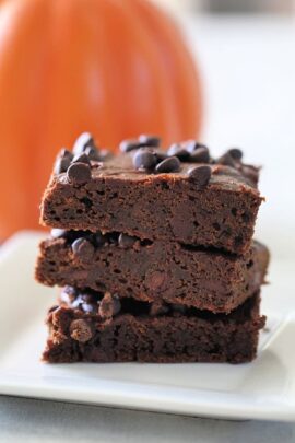 A stack of three pumpkin brownies on a white plate, with an orange pumpkin blurred in the background.