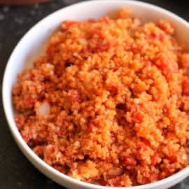 A white bowl filled with a serving of cooked red bulgur wheat on a dark textured surface.