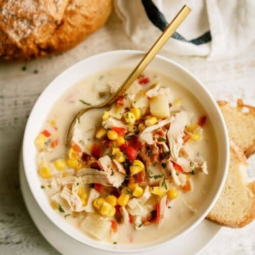 A bowl of creamy chicken and corn chowder garnished with herbs and bacon bits, accompanied by slices of bread on the side.