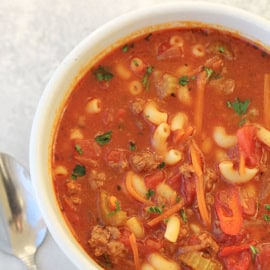 A close-up of a bowl of minestrone soup with visible macaroni, ground meat, chopped vegetables, and a tomato-based broth next to a spoon on a white surface.