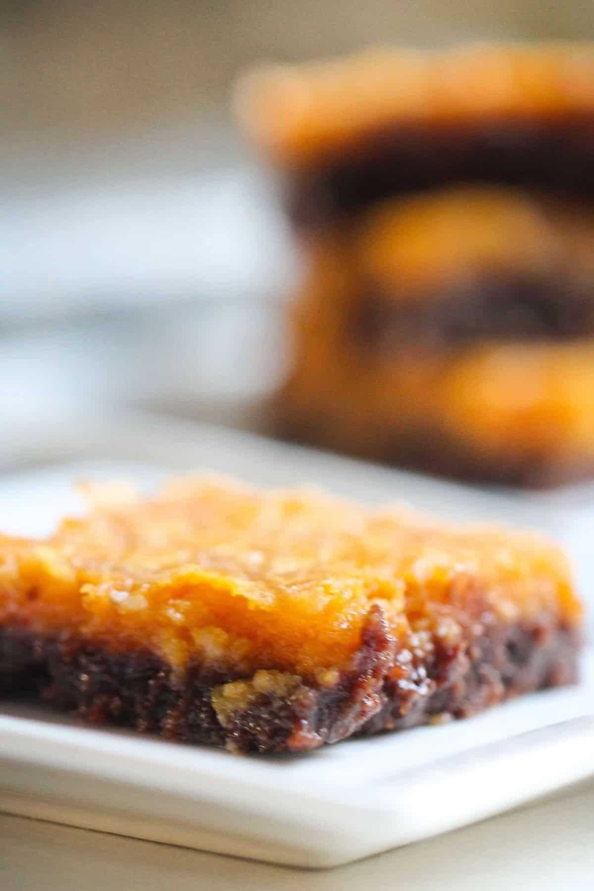 A close-up of a layered dessert bar on a white plate. The bottom layer appears to be chocolate while the top layer is a golden-brown caramel or pumpkin-like topping. Another piece is blurred in the background.