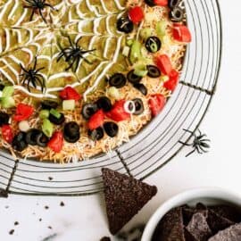 A round seven-layer dip with a spider-web design made of sour cream, topped with tomatoes, olives, and green onions. Plastic spider decorations are around the dip. A bowl of tortilla chips is beside it.