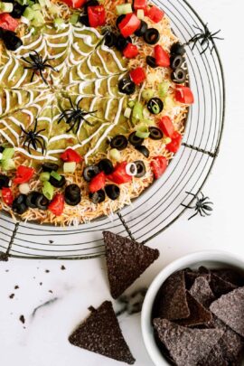 A round seven-layer dip with a spider-web design made of sour cream, topped with tomatoes, olives, and green onions. Plastic spider decorations are around the dip. A bowl of tortilla chips is beside it.