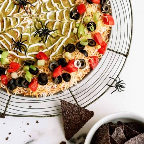 A round seven-layer dip with a spider-web design made of sour cream, topped with tomatoes, olives, and green onions. Plastic spider decorations are around the dip. A bowl of tortilla chips is beside it.