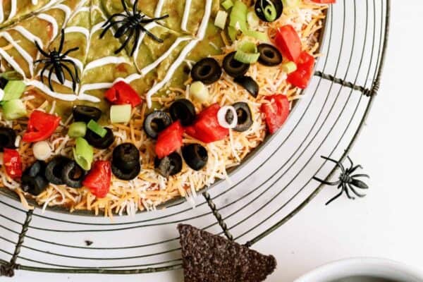 A round seven-layer dip with a spider-web design made of sour cream, topped with tomatoes, olives, and green onions. Plastic spider decorations are around the dip. A bowl of tortilla chips is beside it.