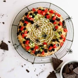 A round platter of layered dip decorated with a spider web-like topping and plastic spiders. Surrounding the platter are scattered chips and a bowl of tortilla chips.