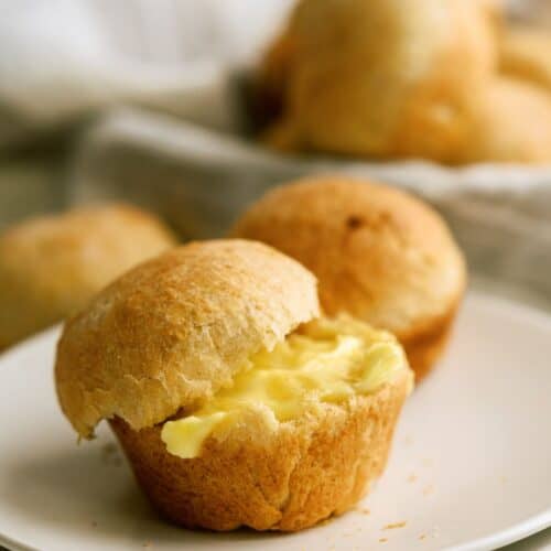 Two buttery rolls are placed on a white plate, one of which is split open, showing a generous amount of butter inside. More rolls are blurred in the background.