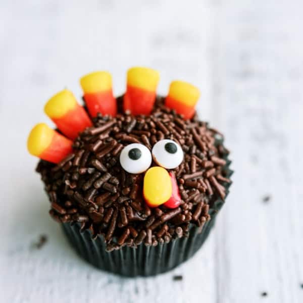 A cupcake decorated with chocolate sprinkles, candy corn arranged as feathers, candy eyes, and a yellow and red nose shaped like a turkey.