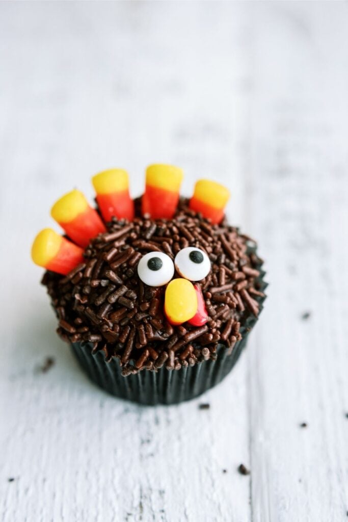 A cupcake decorated with chocolate sprinkles, candy corn arranged as feathers, candy eyes, and a yellow and red nose shaped like a turkey.