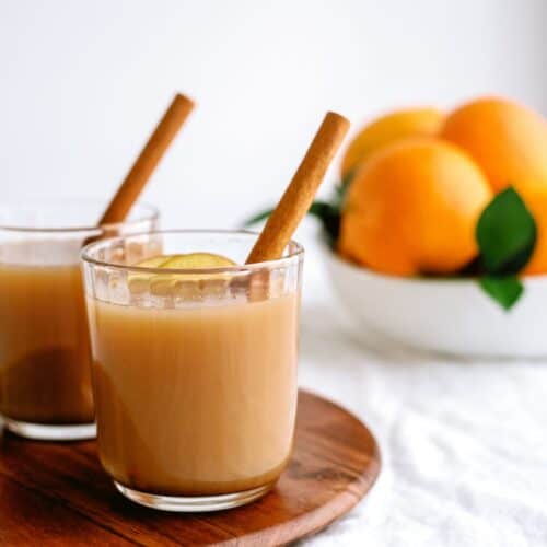 Two glasses of brown liquid with cinnamon sticks and a slice of lemon on a wooden tray; a bowl of oranges in the background.